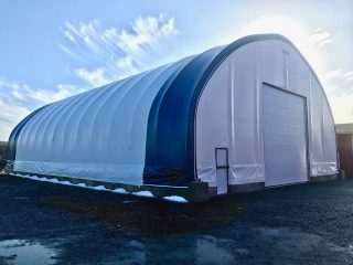 Large white and blue fabric-covered storage building with an arched roof, concrete base, and a tall roll-up door, set on a gravel lot under a partly cloudy sky.