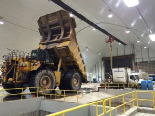 A large yellow dump truck with its bed raised is inside an industrial building, near a worker on a raised platform and various equipment. Yellow railings border the area.