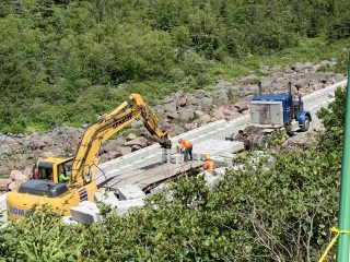 A yellow excavator and a blue truck are parked beside a concrete road under construction. Workers in orange vests are on site, surrounded by rocks, greenery, and forested hills in the background.