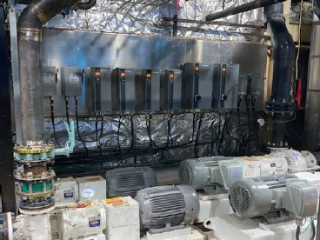 Several industrial electric motors and pumps are mounted on a platform, with large black and silver pipes connected above. Behind them, control panels with multiple switches and wiring are attached to a reflective metallic wall.