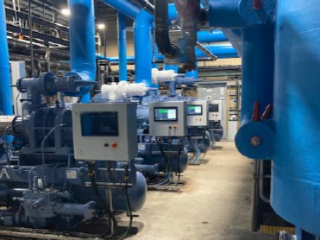 Rows of large blue industrial machines and control panels are positioned in a spacious, brightly lit facility with concrete floors and exposed pipes overhead.