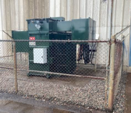 A large green electrical transformer is enclosed by a rusty chain-link fence, situated on gravel next to a beige industrial building with stained walls. The ground outside the fence appears wet.