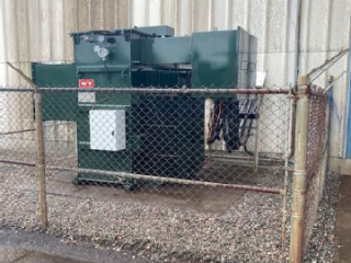 A large green electrical transformer is enclosed by a rusty chain-link fence, situated on gravel next to a beige industrial building with stained walls. The ground outside the fence appears wet.