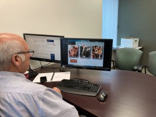 A man with gray hair and glasses sits at a desk using a dual-monitor setup; one screen displays text, and the other shows images of motorcycles and the word “INDIAN.” A keyboard, mouse, and notepad are on the desk.