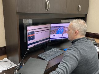 A man with short blond hair sits at a desk using two computer monitors, one displaying a website and the other showing a blue abstract image. A keyboard, mouse, and closed laptop are also on the desk.