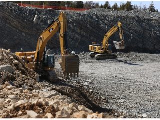 Two yellow excavators are digging and moving rocks in a large, open quarry with rocky walls and scattered debris. Trees and an orange safety barrier are visible in the background.