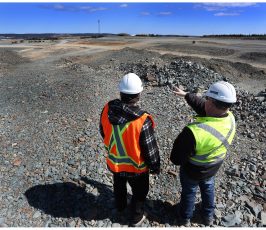 Two workers wearing safety vests and hard hats stand on rocky ground at a construction or mining site, with one pointing towards the distance under a blue sky.
