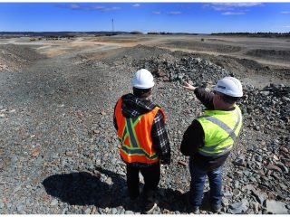 Two workers wearing safety vests and hard hats stand on rocky ground at a construction or mining site, with one pointing towards the distance under a blue sky.
