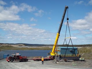 A yellow crane lifts a large blue container onto a red flatbed truck near a lake, while a worker in a safety vest observes on a gravel road under a partly cloudy sky.