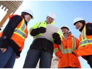 Four construction workers in safety gear and hard hats stand outdoors, looking at a document and smiling. A large piece of construction equipment is visible in the background against a blue sky.