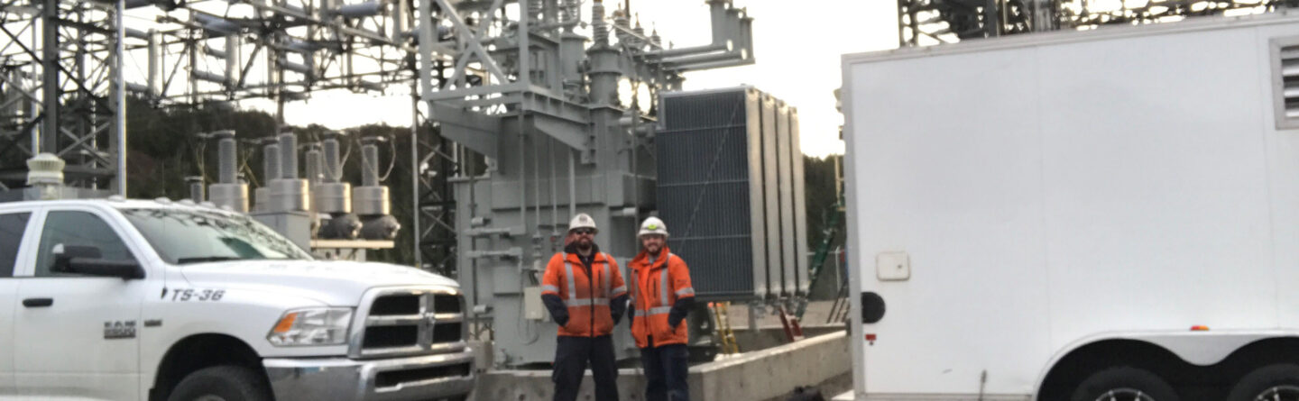 Two workers in orange safety jackets and helmets stand near electrical equipment at a substation, between a white truck and a white trailer, surrounded by metal structures and wires.
