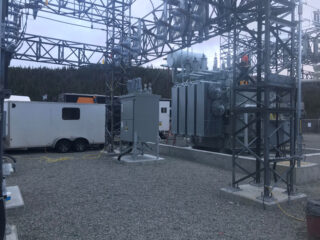 Electrical substation with large transformers, metal structures, and power lines. Two white utility trailers are parked nearby. The area is covered with gravel, and trees are visible in the background.