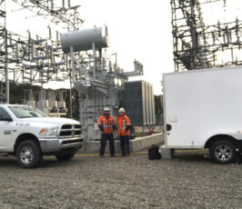 Two workers in orange safety jackets and helmets stand near electrical equipment at a substation, between a white truck and a white trailer, surrounded by metal structures and wires.