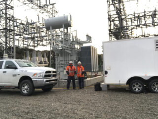 Two workers in orange safety jackets and helmets stand near electrical equipment at a substation, between a white truck and a white trailer, surrounded by metal structures and wires.