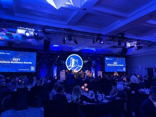 A banquet hall filled with people seated at round tables during the 2021 Business Resilience Awards. Blue lighting highlights the stage, where award presentations and speeches are taking place. Two large screens display the event name.
