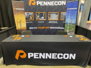 A PENNECON booth at an event, featuring a black tablecloth with the company logo, orange pens, and brochures neatly placed on the table, with a branded backdrop displaying images of engineering projects.