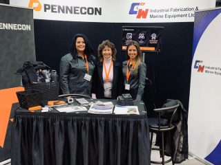 Three women stand smiling behind a booth at a trade show. The booth displays the logos for Pennecon and C&W Industrial Fabrication & Marine Equipment Ltd., with pamphlets and promotional materials on the table.