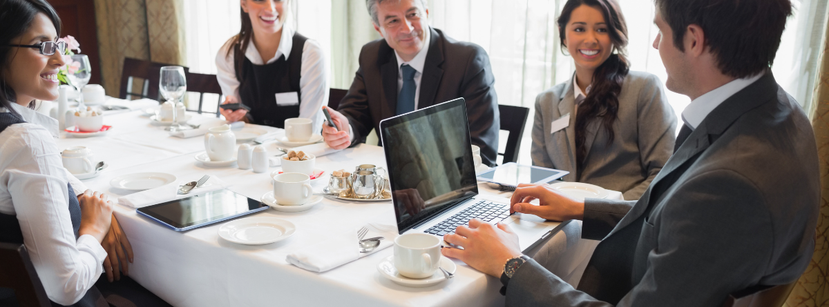 Five business professionals sit around a table in a restaurant, having a meeting. They are smiling, with laptops, tablets, and coffee cups on the table, suggesting a collaborative and positive atmosphere.