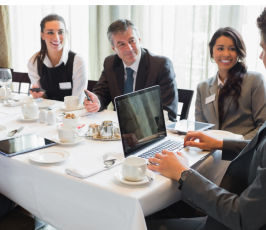 Five business professionals sit around a table in a restaurant, having a meeting. They are smiling, with laptops, tablets, and coffee cups on the table, suggesting a collaborative and positive atmosphere.