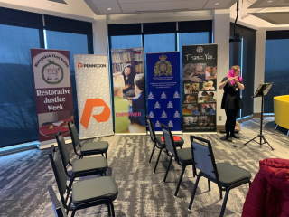 A small event setup with five banners behind rows of empty chairs; a woman plays a flute on the right. The banners promote Restorative Justice Week, Pennecon, RCMP, and an animal welfare group.