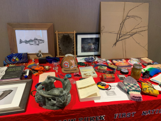 A tabletop covered with various Indigenous art pieces, beadwork, crafts, books, knitted items, framed artwork, a jar of preserves, and a large cardboard with branches. A red cloth reads 