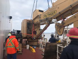 Workers in safety gear operate and supervise a large PALFINGER crane on a ship's deck near the water, with construction materials and equipment around them under a cloudy sky.