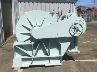 A large industrial winch painted light grey sits on a concrete surface outside a metal building, with a blue sky and some scattered clouds in the background.