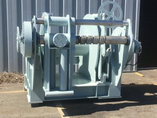 A large industrial winch with a metal drum and gear mechanisms sits on a paved outdoor surface in front of a corrugated metal building.