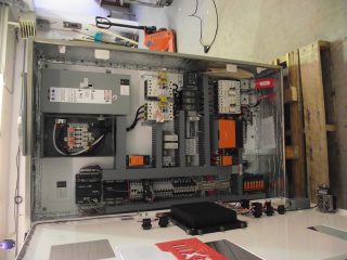 Open industrial electrical control panel with wires, circuit breakers, relays, and various electronic components mounted inside a metal cabinet, situated on the floor next to wooden pallets and some cables.