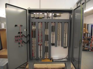 An open industrial electrical control panel with organized wiring, relays, terminals, and circuit boards mounted inside a metal cabinet; the panel doors are open, revealing all internal components.