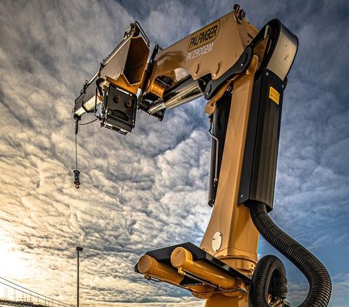 A large yellow industrial robotic arm extends upwards outdoors under a dramatic cloudy sky, with a bridge and water visible in the background.