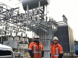 Two workers in orange high-visibility jackets and hard hats stand in front of large electrical equipment at an outdoor power substation. Metal structures and wires are visible in the background.