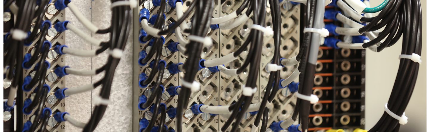 Close-up of a network server panel with multiple black cables connected to labeled ports, secured with white cable ties, showing organized wiring in a data center or telecom setup.
