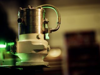 A close-up of a cylindrical metal industrial component with attached pipes and bolts, illuminated by green light, set against a blurred background.