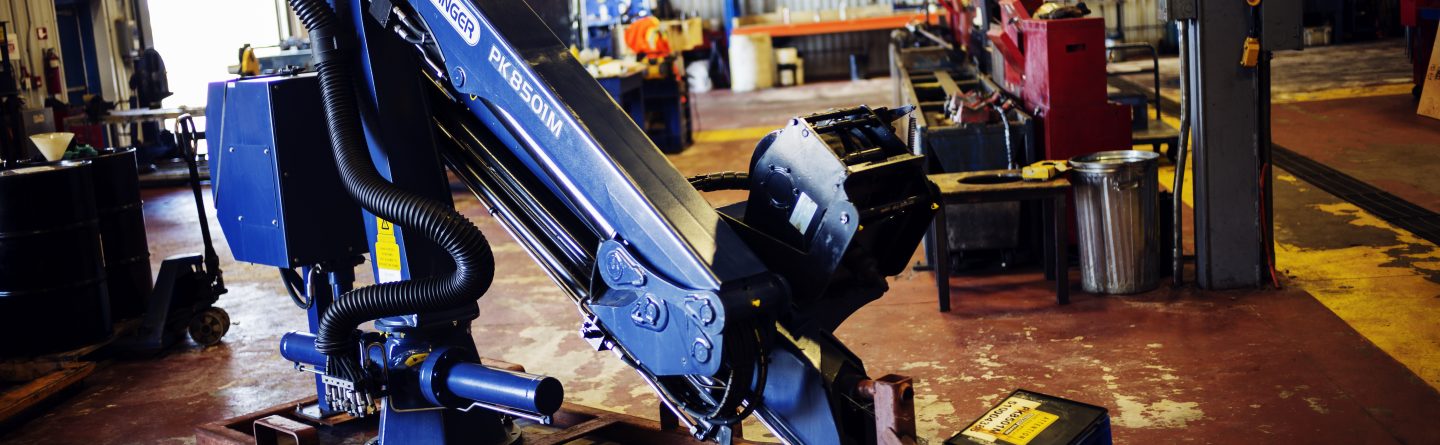 A robotic welding arm, labeled "FANUC ARC MATE 120i," is set up inside an industrial workshop with shelves, tools, and machinery in the background. The floor is stained, and lighting is natural from windows.