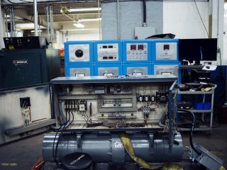 An industrial setting with an open control panel showing wiring and components, blue electrical cabinets with gauges and switches in the background, a computer monitor, and various equipment and tools nearby.