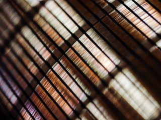 Close-up of a bamboo or wooden window blind, with sunlight filtering through the slats, creating a warm, patterned play of light and shadow.