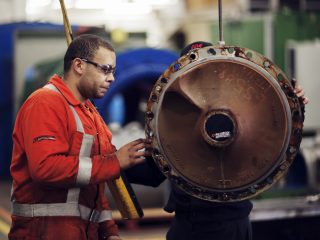 Two workers in a factory, one in an orange jumpsuit and safety glasses, examine a large, round mechanical part suspended in the air. Industrial equipment and machinery are visible in the background.