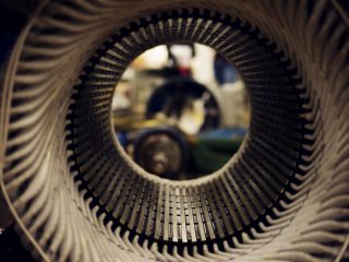 Close-up view through the circular interior of an industrial machine part, possibly a stator or turbine, with metal fins in focus and a blurred workshop background visible through the center opening.