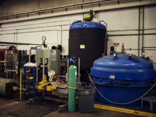 Industrial workshop with large blue machinery, control panels, oxygen tank, tools, and safety barriers; various equipment and cables are arranged around the spacious room with white brick walls.