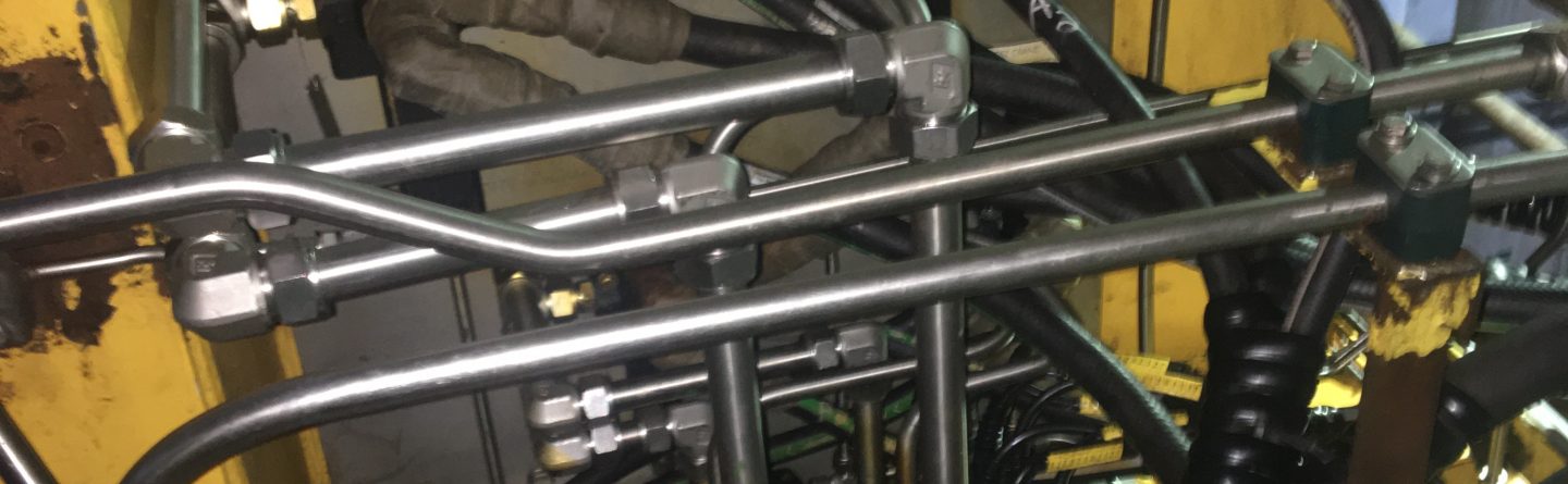 Close-up of industrial machinery showing a yellow metal enclosure with multiple black hoses, silver pipes, and connectors attached, displaying a complex network of hydraulic or pneumatic lines.