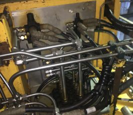 Close-up of industrial machinery showing a yellow metal enclosure with multiple black hoses, silver pipes, and connectors attached, displaying a complex network of hydraulic or pneumatic lines.