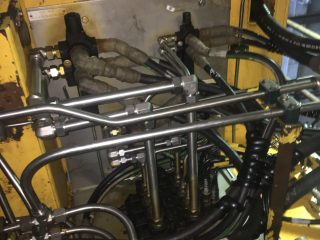 Close-up of industrial machinery showing a yellow metal enclosure with multiple black hoses, silver pipes, and connectors attached, displaying a complex network of hydraulic or pneumatic lines.