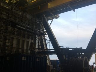 An industrial interior shows scaffolding, metal beams, a large yellow overhead crane, and blue containers, with a view of the sky and horizon visible through a large open section of the wall.