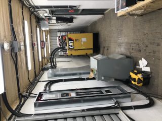 An industrial room with a large yellow CAT generator, electrical panels, wiring, a grey utility box, and various equipment on a concrete floor. The walls are lined with cables and insulation.
