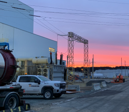 A construction site at sunset, featuring a white PennEcon truck, industrial equipment, and large building structures. The sky is vibrant with pink and purple hues.