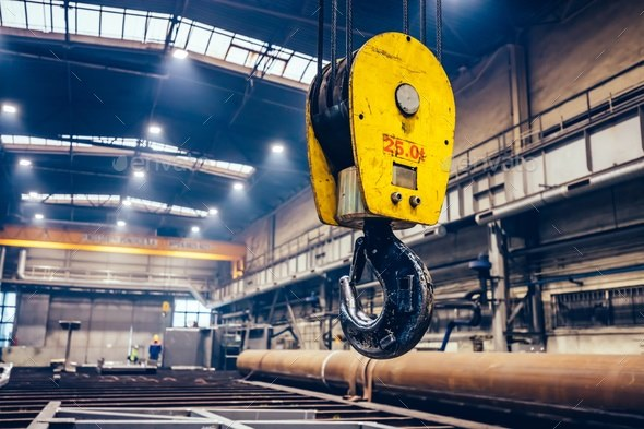 Large yellow industrial crane hook hanging in a spacious factory or warehouse, with metal pipes and beams below, workers visible in the background, and light streaming through high windows.