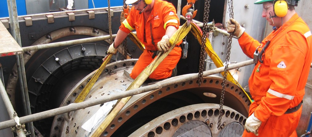 Two workers in orange safety gear and helmets use straps and a crane to move a large industrial turbine part inside a factory or plant, surrounded by machinery and metal components.