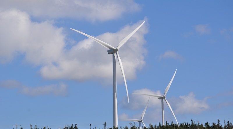 Two large white wind turbines stand above a forested landscape under a blue sky with scattered clouds.
