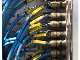 Close-up of multiple blue cables connected to brass fittings on a metal panel, each cable labeled with yellow tags featuring black numbers and letters. The cables are bundled and neatly organized.